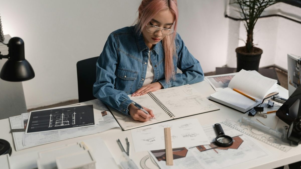 A person sketching architectural plans at a desk covered with drawings